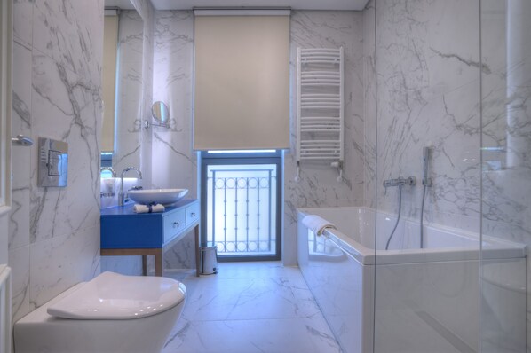 A modern bathroom with a marble floor, a white bathtub, a glass-enclosed shower, and a blue vanity with a sink. - Hotel Majestic (Budva)