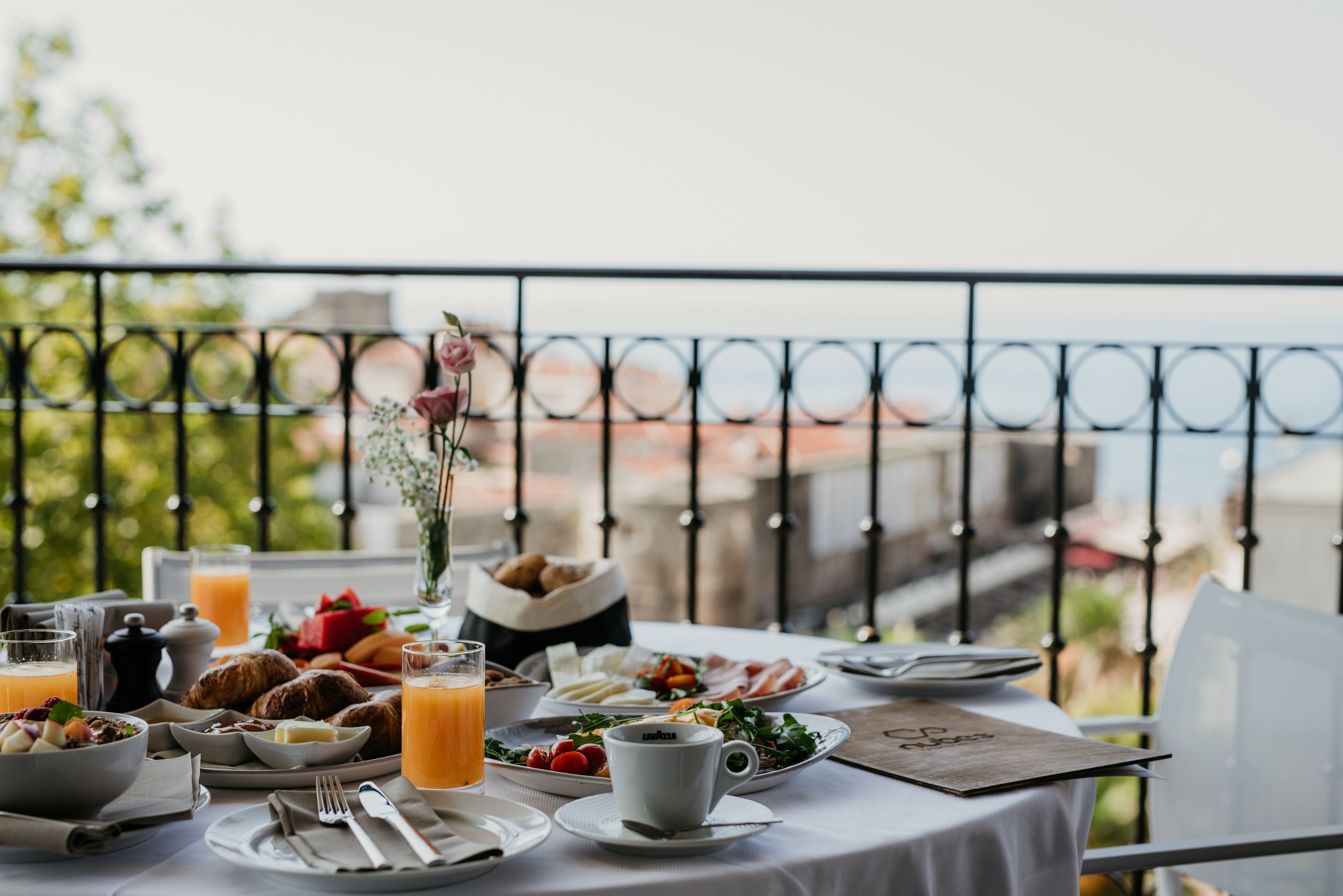 Una mesa de desayuno con cruasanes, fruta y zumo de naranja en un balcón con vistas a la ciudad.