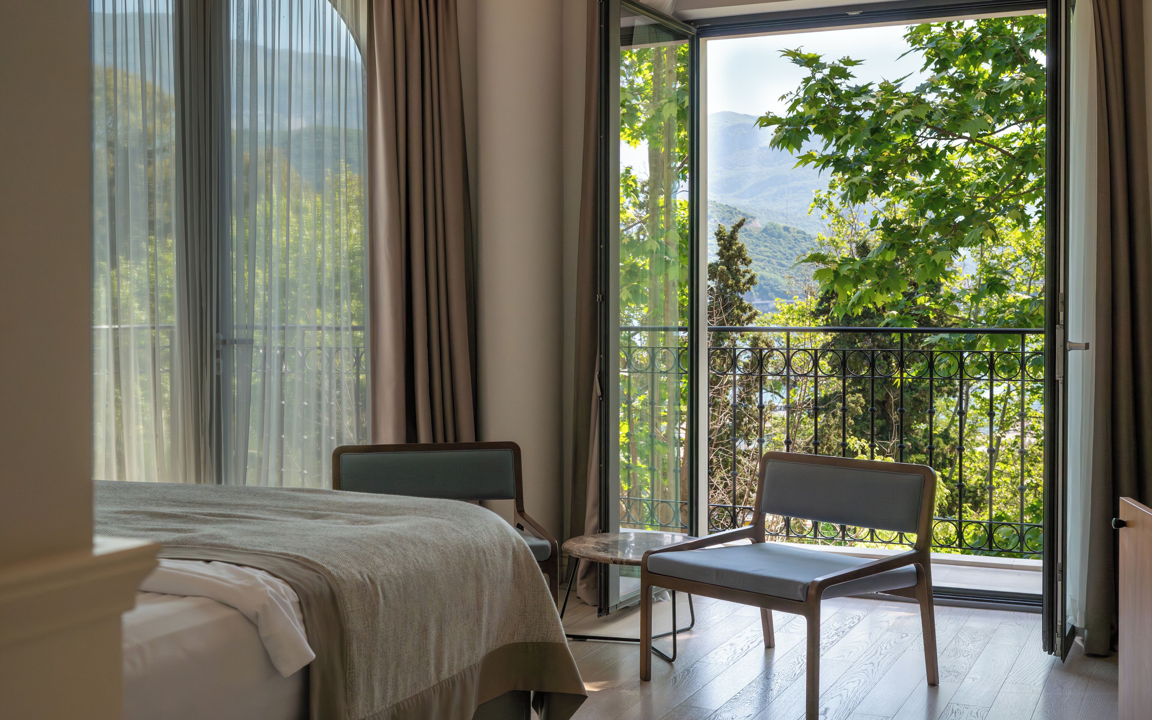 A bedroom with a bed, a chair, a table, and a balcony with a view of trees and mountains.