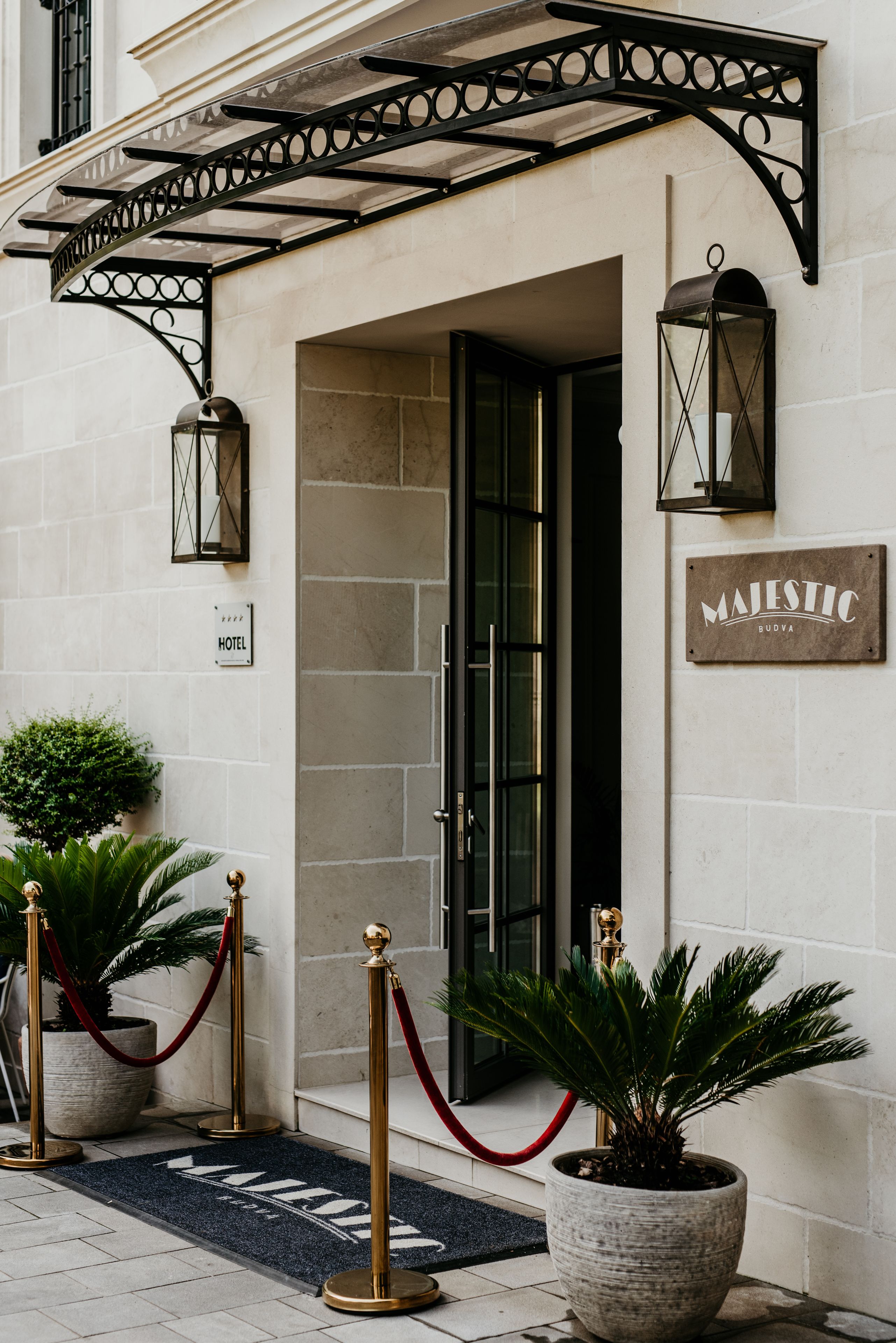 A hotel entrance with a wrought iron canopy, a sign reading "MAJESTIC BUDVA," and a doormat with the hotel's name.