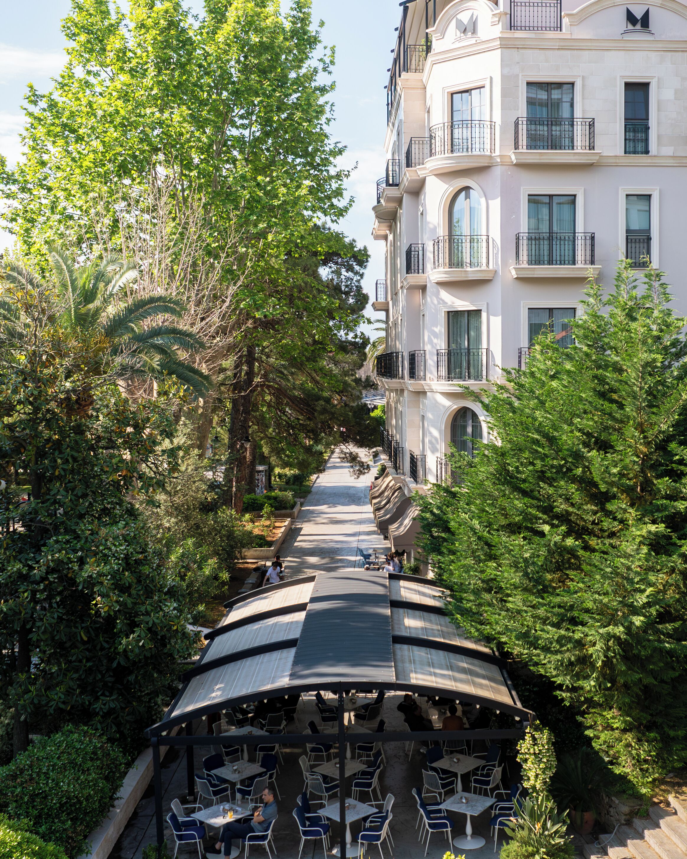 Una calle arbolada con mesas al aire libre y un edificio de varios pisos con balcones.