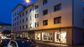 Um hotel com vários andares e um letreiro chamativo com a palavra 'HOTEL', uma loja boutique no térreo e uma vista da rua com carros estacionados.