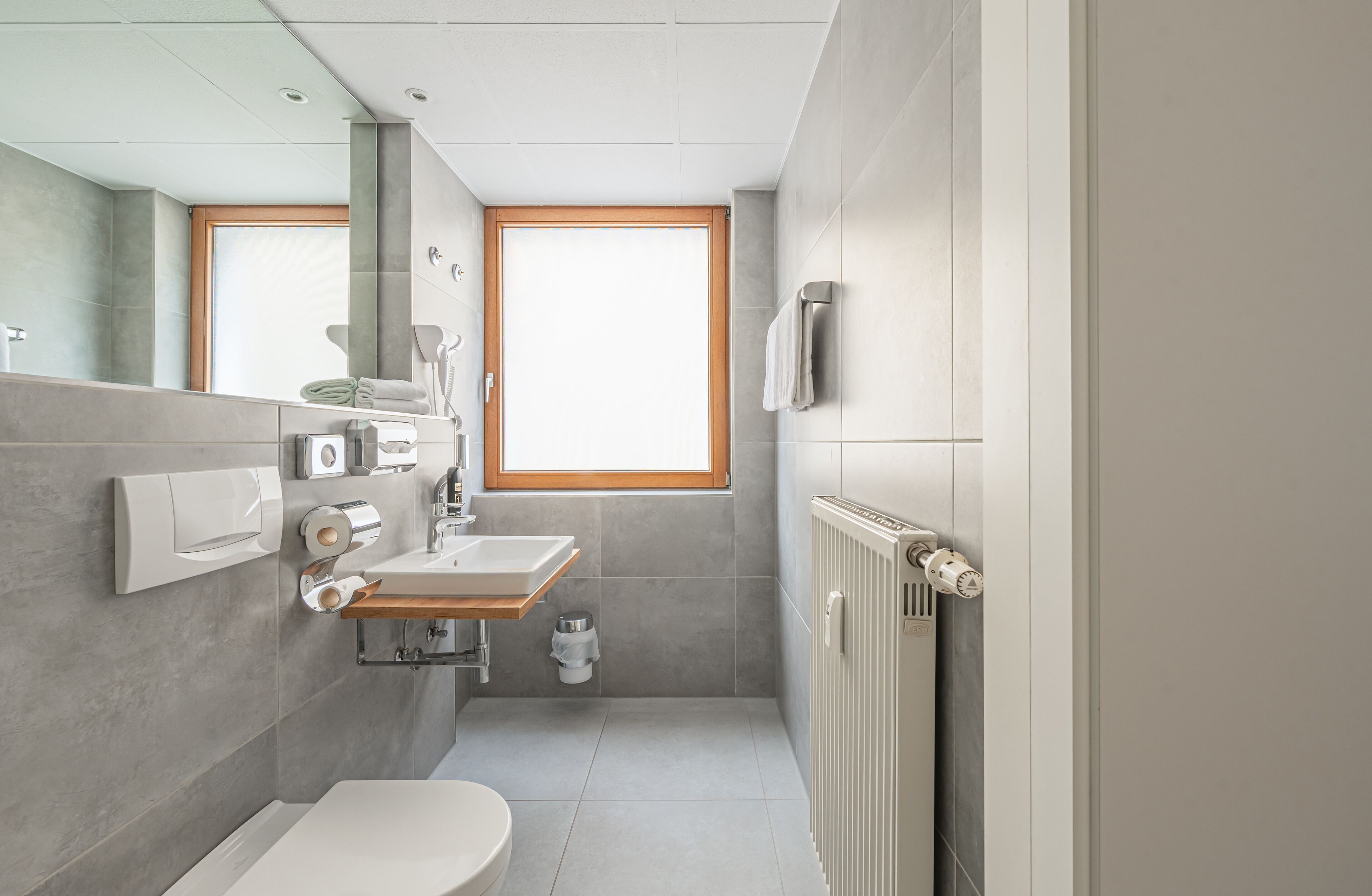 A modern bathroom with a toilet, sink, and mirror. There is a window providing natural light and a towel rack on the wall.