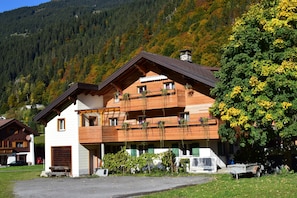 A two-story wooden house with a balcony, surrounded by a lush green landscape and a forested mountain in the background. - Ferienwohnung Birgit (Sankt Gallenkirch)