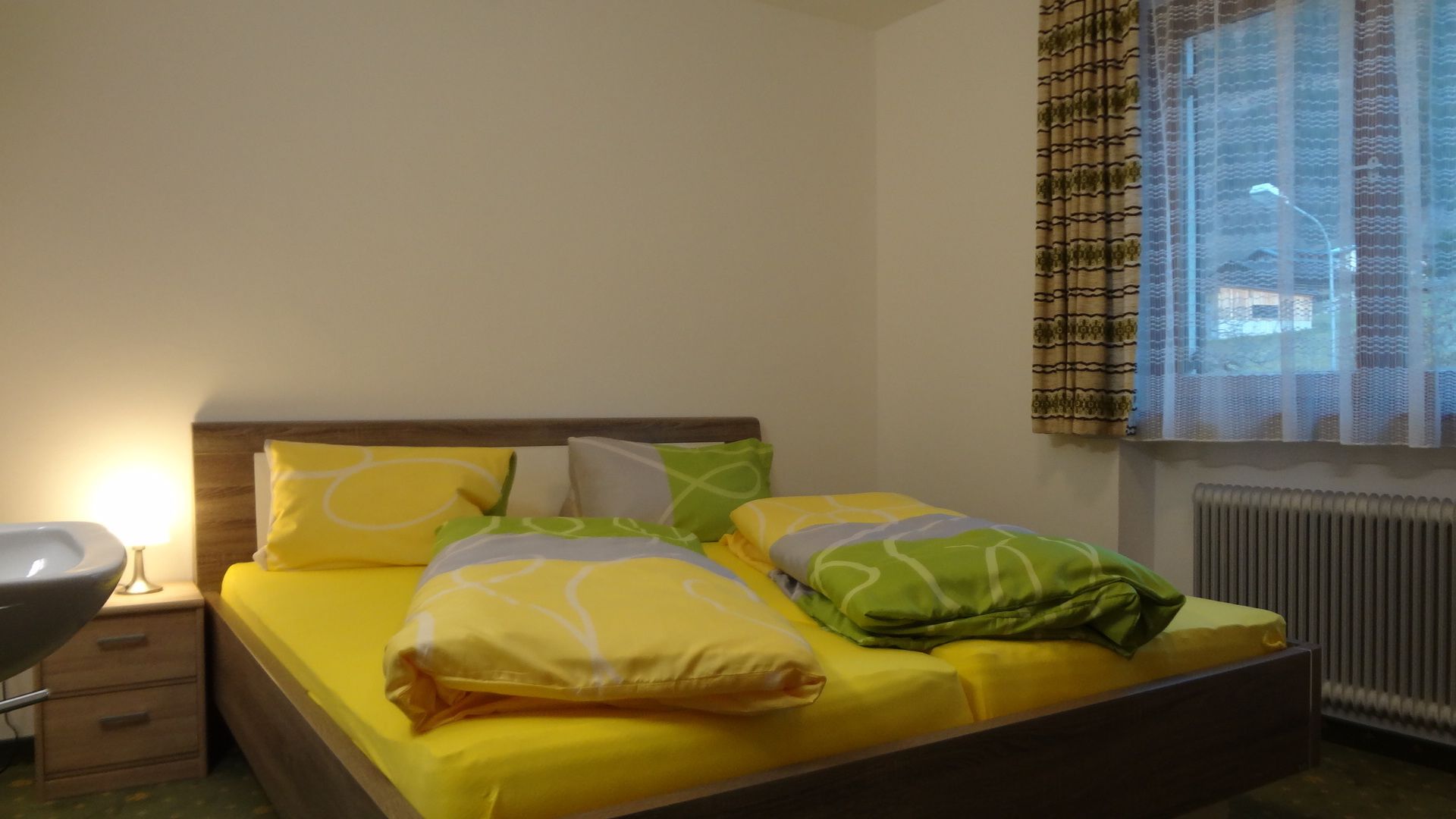 Een bed met geel beddengoed en groene kussens, een nachtkastje met een lamp, en een raam met gordijnen met een patroon.