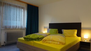 A hotel room with a bed, a nightstand, a lamp, a window with curtains, and a radiator. - Ferienwohnung Alexander (Sankt Gallenkirch)