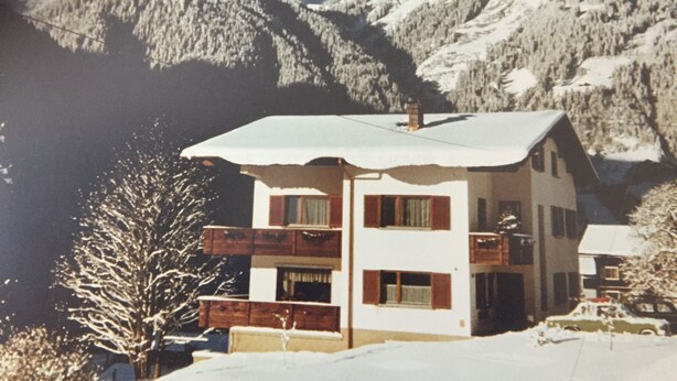 A two-story chalet with a balcony, set against a backdrop of snow-covered mountains and a lake. - Ferienwohnung Alexander (Sankt Gallenkirch)