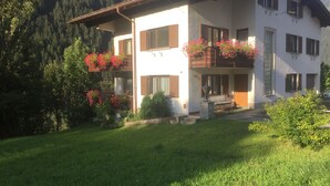 A two-story house with a balcony, surrounded by greenery and a mountainous backdrop. - Ferienwohnung Alexander (Sankt Gallenkirch)