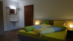 A bedroom with a bed, a bedside table with a lamp, a wooden door, a wall-mounted sink, and a mirror. - Ferienwohnung Alexander (Sankt Gallenkirch)