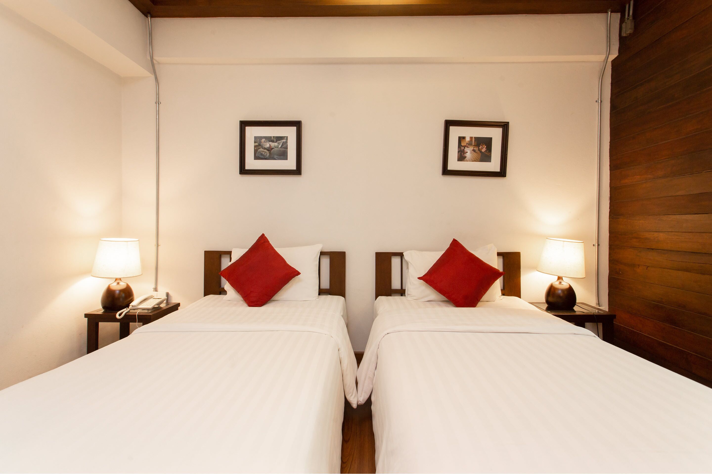 A hotel room with two beds, each with a red pillow and a bedside lamp. There are framed pictures on the wall above the beds.