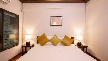 Une chambre d’hôtel avec un grand lit, trois oreillers, deux tables de chevet avec des lampes, une œuvre d’art encadrée au mur et une fenêtre avec des stores.