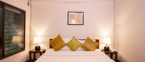 A hotel room with a large bed, three pillows, two bedside tables with lamps, a framed artwork on the wall, and a window with blinds.