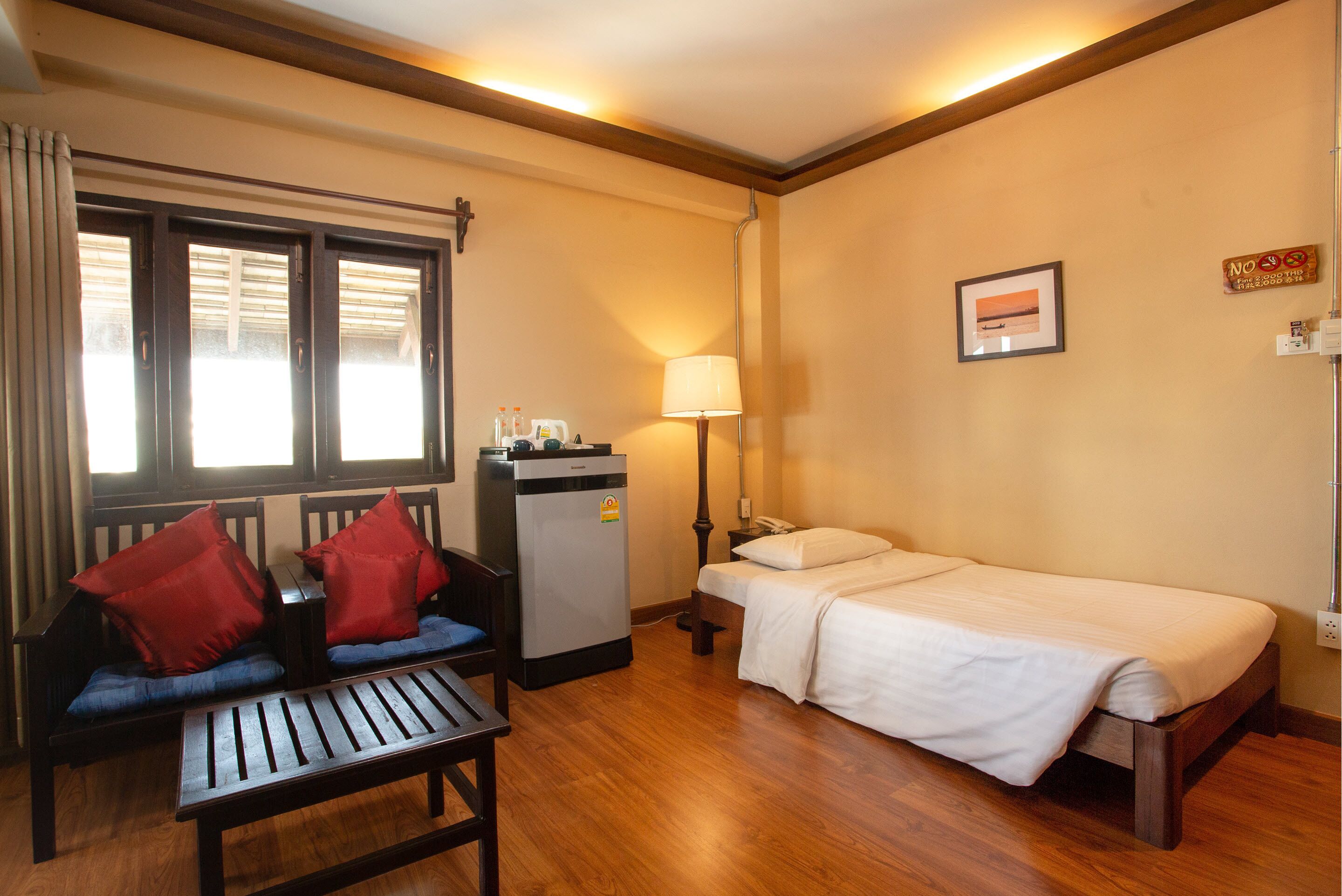 A compact hotel room with a single bed, a small desk with a refrigerator, a chair with red cushions, and a window with blinds.