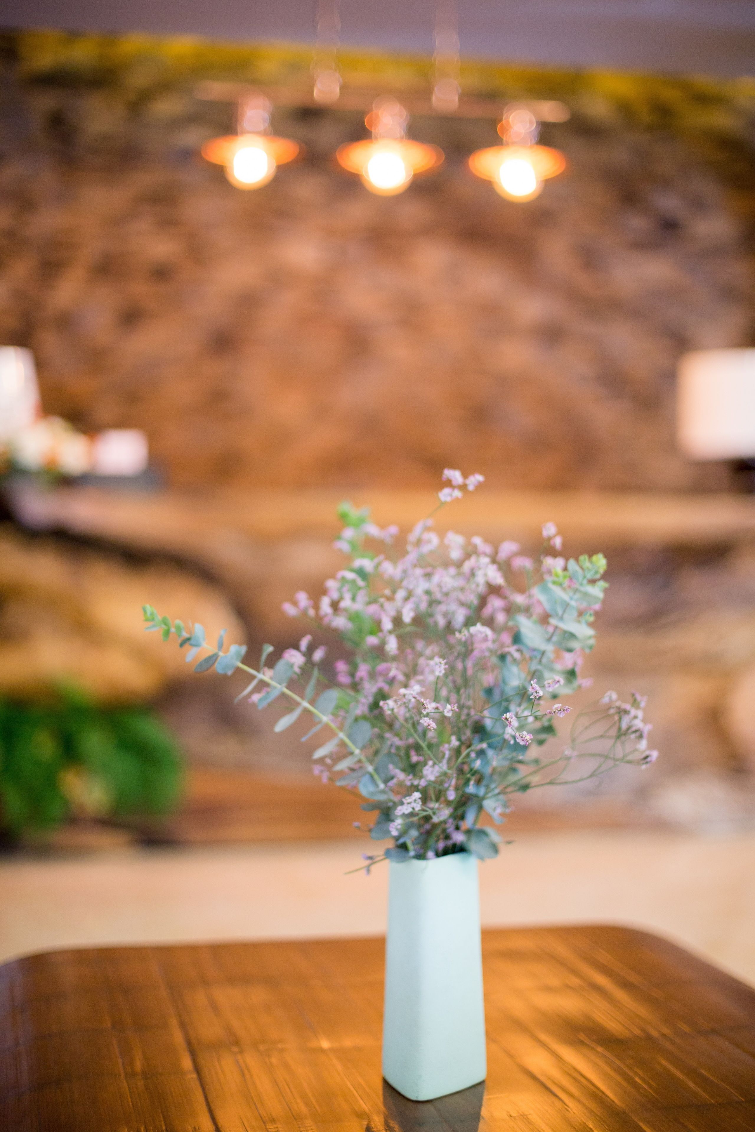 Een witte vaas met bloemen op een houten tafel in een kamer met een stenen muur en hangende lampen.