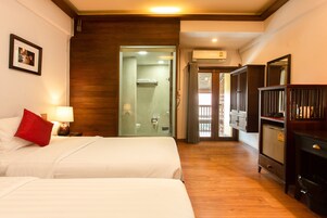 Habitación de hotel con dos camas, suelo de madera, armario y baño con puerta de vidrio.