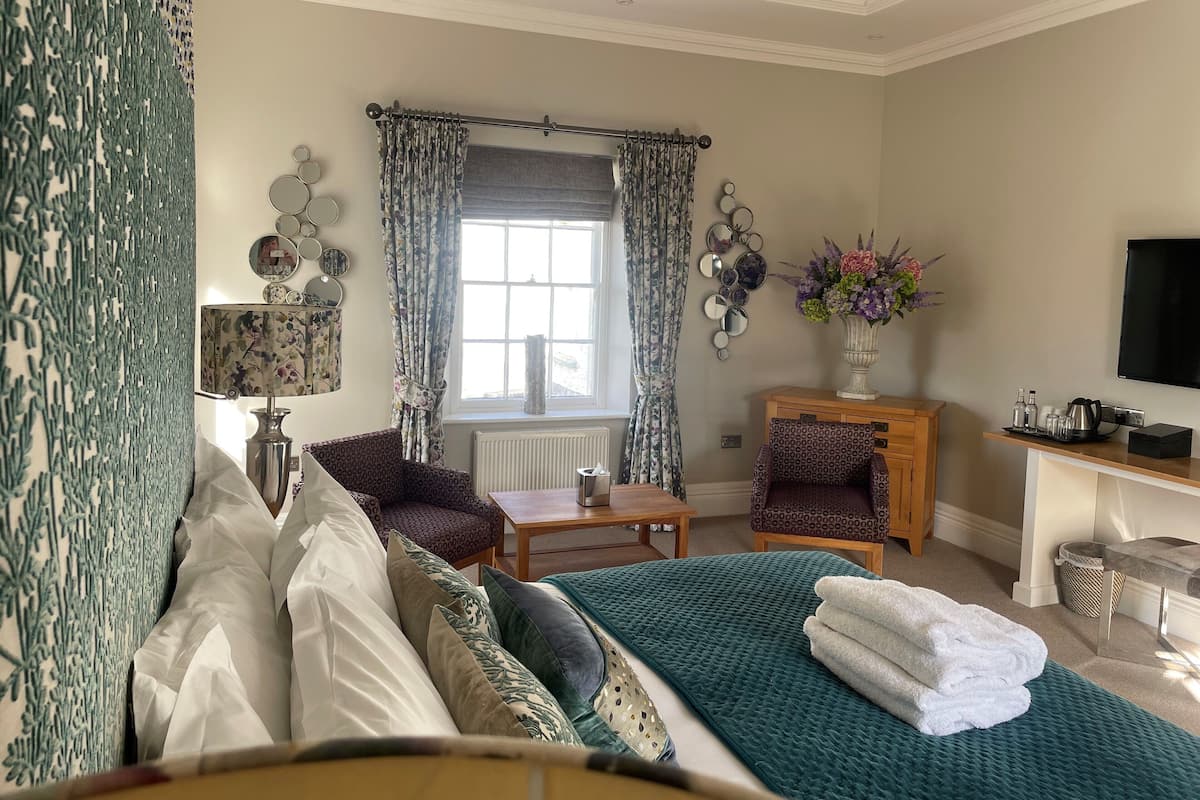 Une chambre d’hôtel comprenant un lit, deux fauteuils, une petite table, un bureau avec une télévision, une fenêtre avec des rideaux et un vase de fleurs.