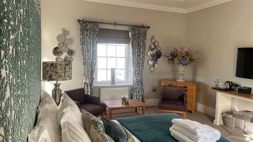 A hotel room with a bed, two armchairs, a small table, a desk with a TV, a window with curtains, and a vase with flowers.