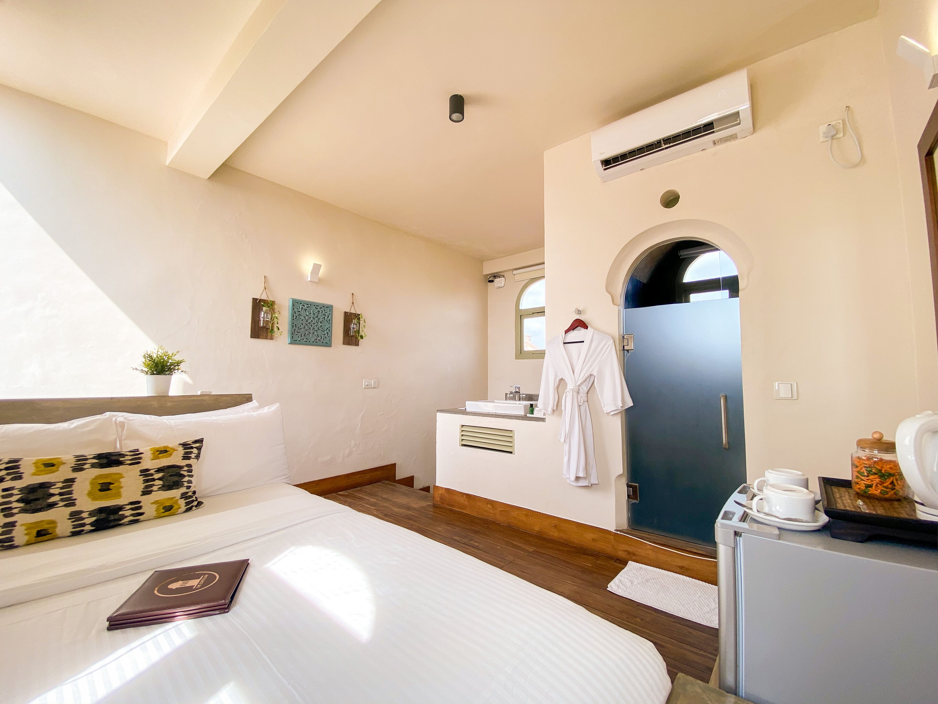 A hotel room with a bed, a bedside table, a small desk, a mirror, a bathroom with a sink, and an air conditioning unit.