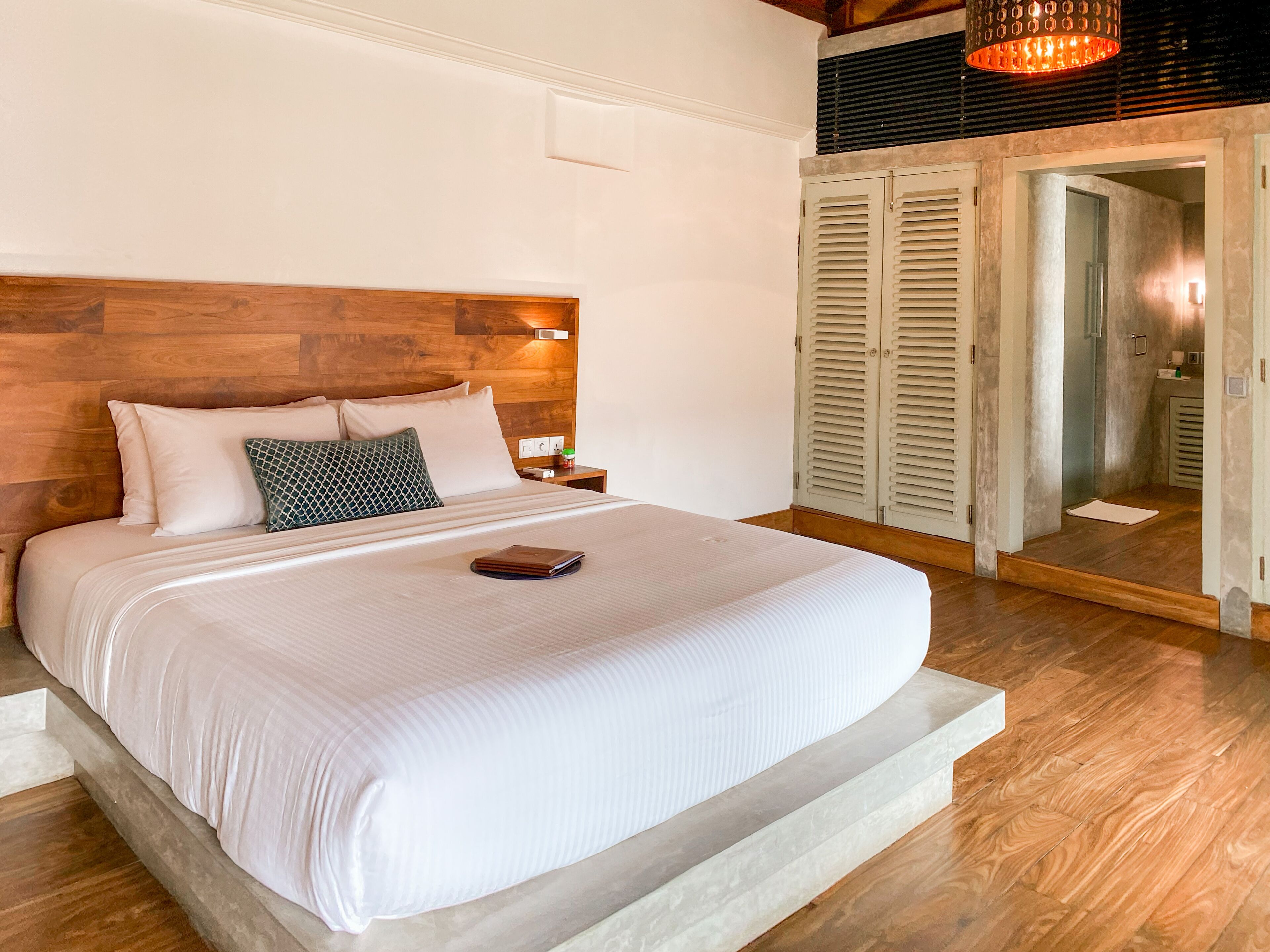 A neatly made bed with a wooden headboard, a bedside table with a lamp, and a view of a bathroom through a door.