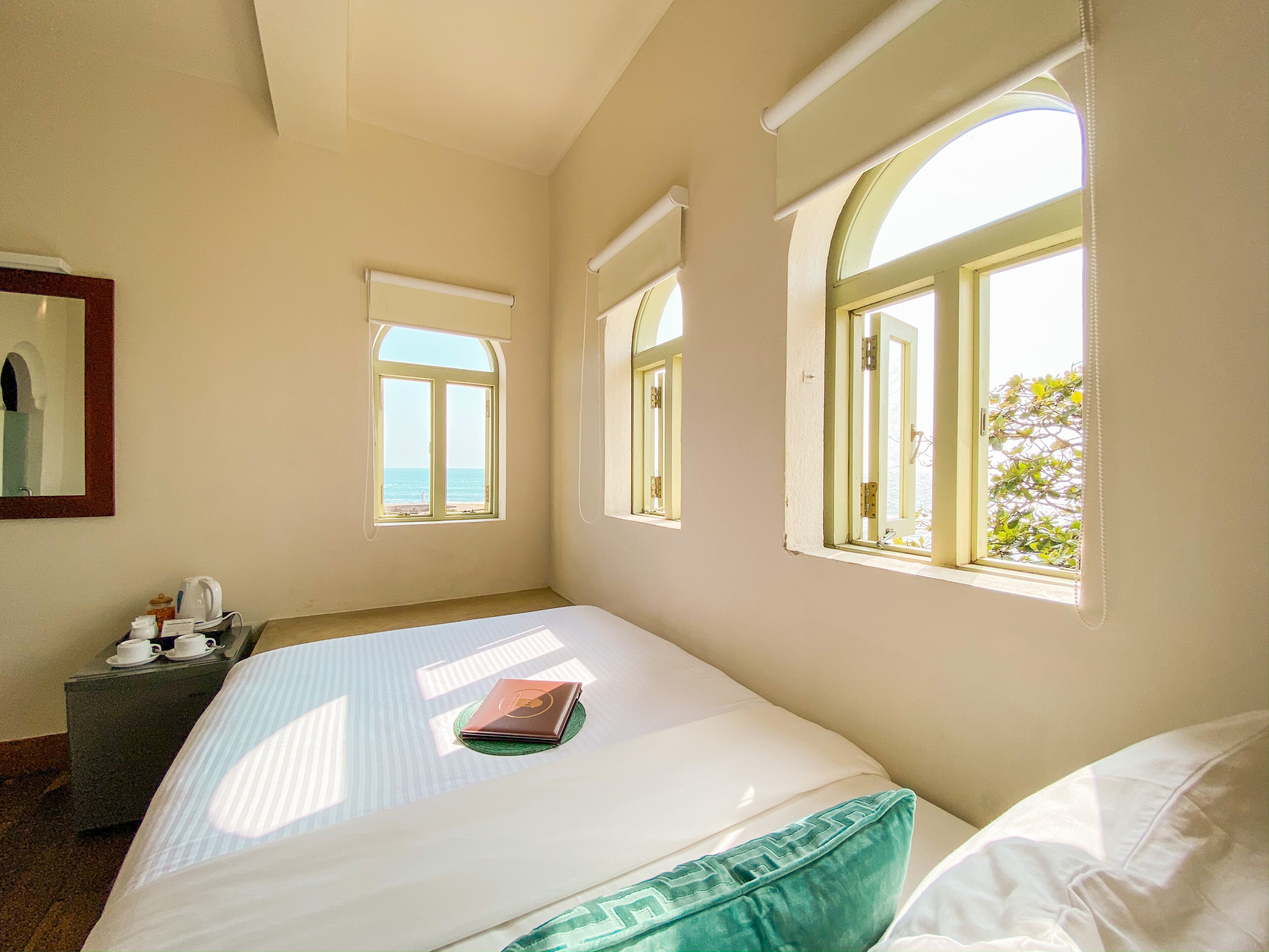 A hotel room with a bed, a bedside table with a lamp, and a window with a view of the sea.