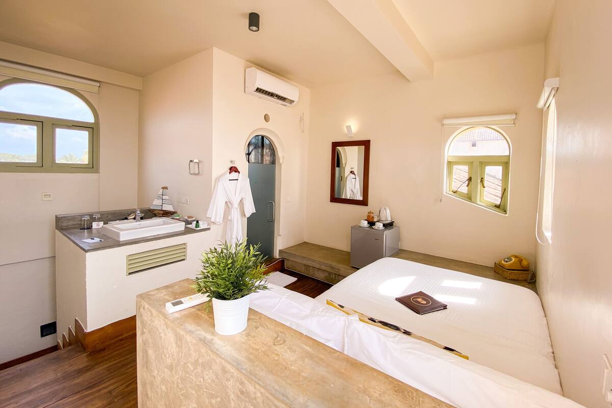 Una camera d'albergo con un letto, un comodino con una pianta, un bagno con un lavandino e uno specchio, e una finestra vista.