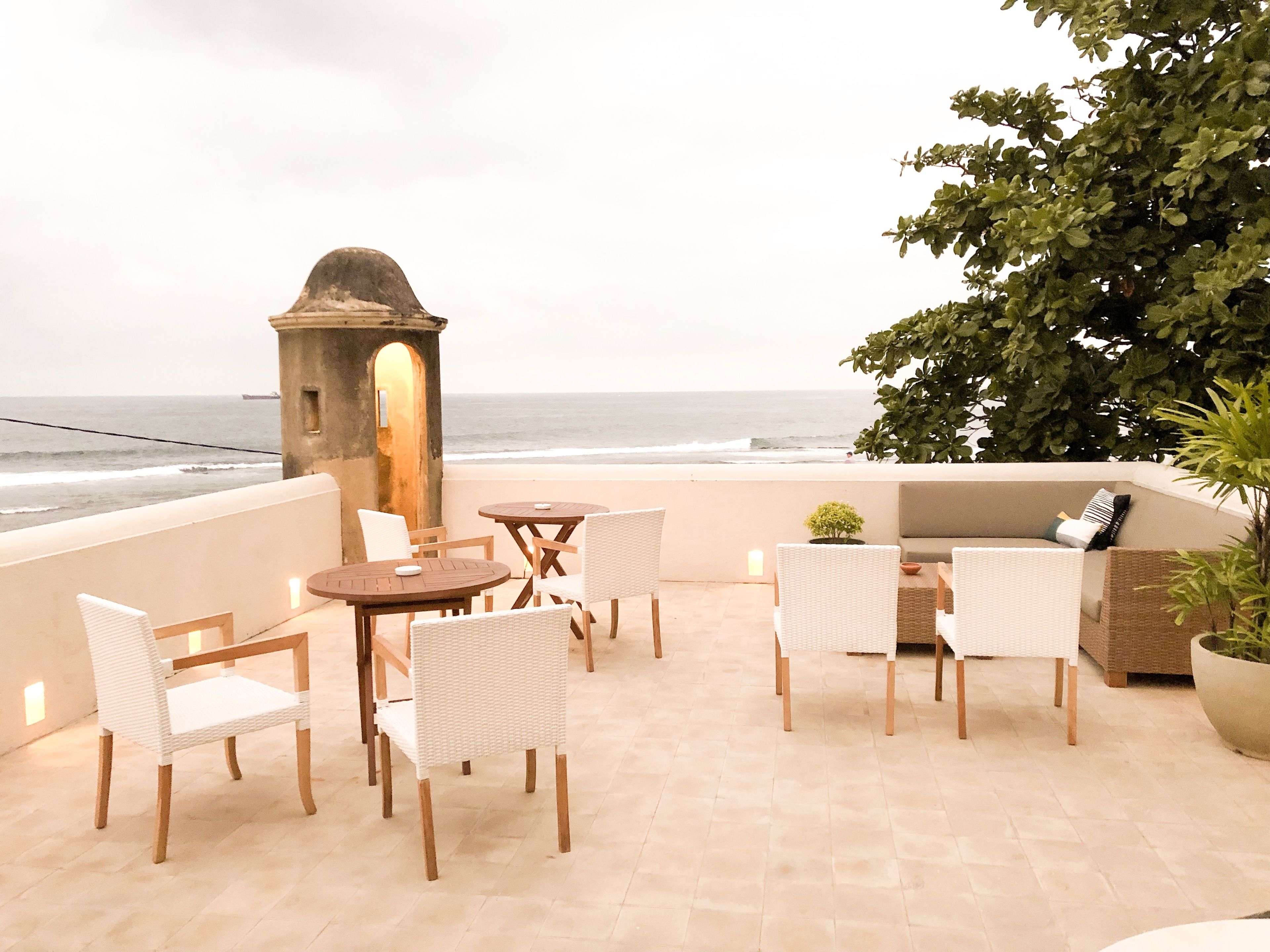 A terrace with a view of the ocean, featuring a stone archway, wicker furniture, and a potted plant.