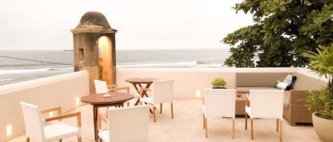 A terrace with a view of the ocean, featuring a stone archway, wicker furniture, and a potted plant.