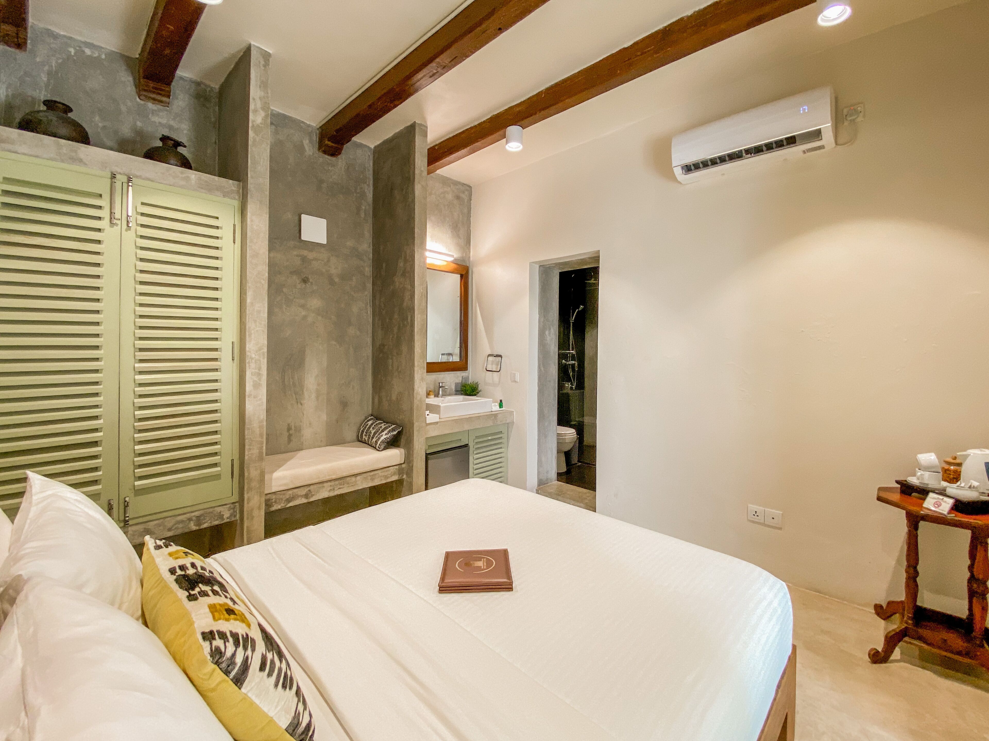 Un dormitorio con cama, mesita de noche, espejo, y un baño visible a través de una puerta abierta, además de una viga de madera en el techo.