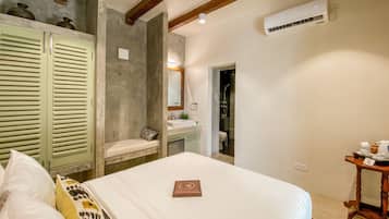 Un dormitorio con cama, mesita de noche, espejo, y un baño visible a través de una puerta abierta, además de una viga de madera en el techo.