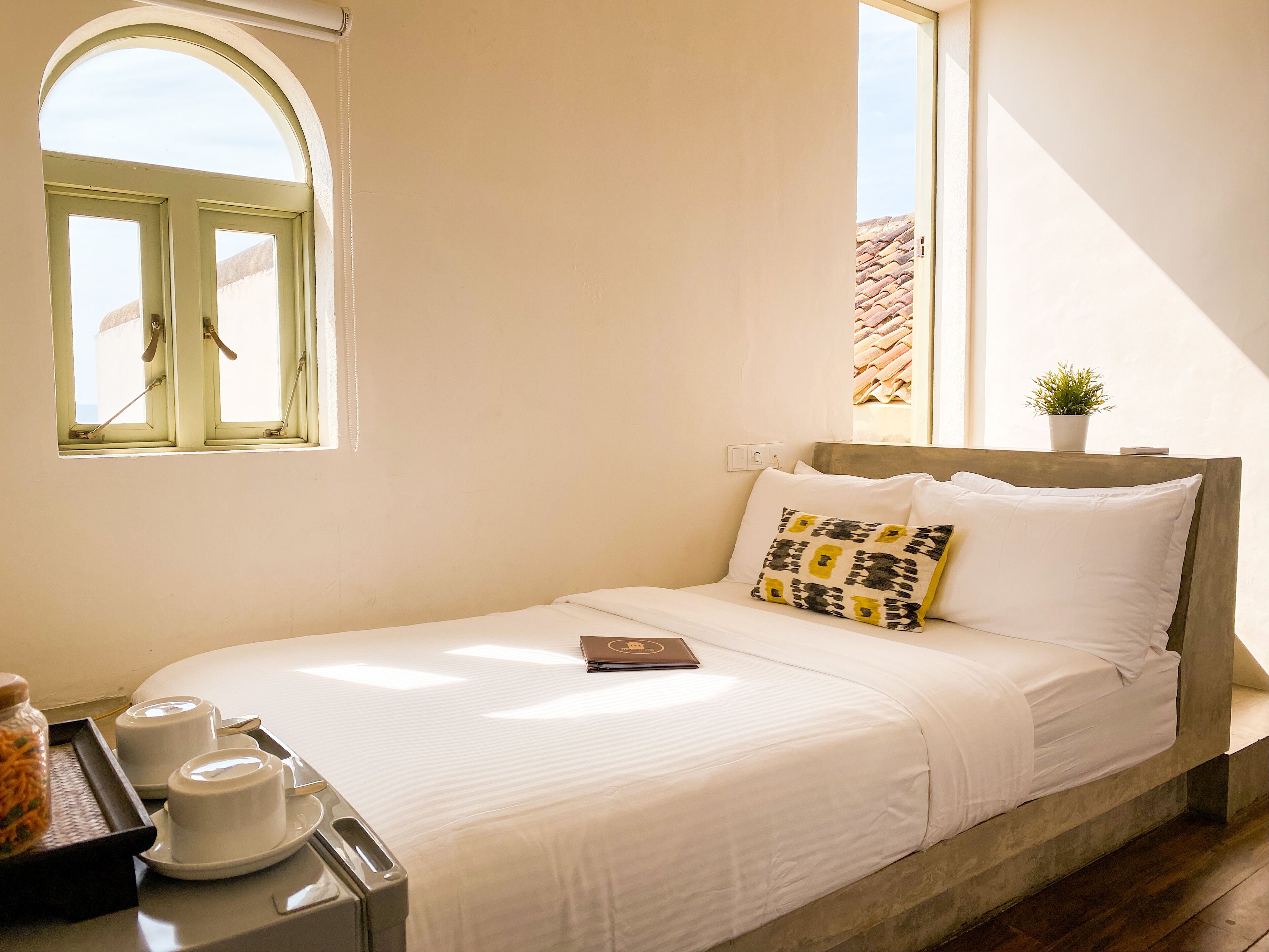 A bed with white bedding, a pillow with a geometric pattern, a small potted plant, and a window with a view of a building.