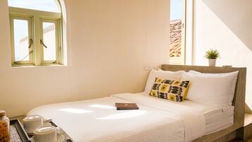 A bed with white bedding, a pillow with a geometric pattern, a small potted plant, and a window with a view of a building.