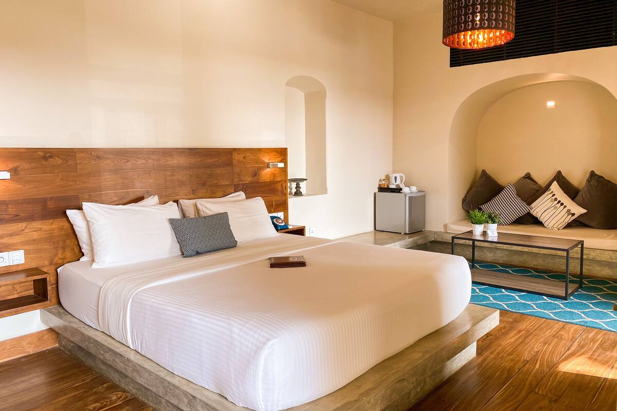 Una camera d'albergo moderna con un letto grande, una testiera in legno, un piccolo angolo relax con cuscini e un tavolino da caffè.