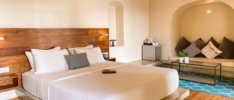 A modern hotel room with a large bed, a wooden headboard, a small seating area with cushions, and a coffee table.