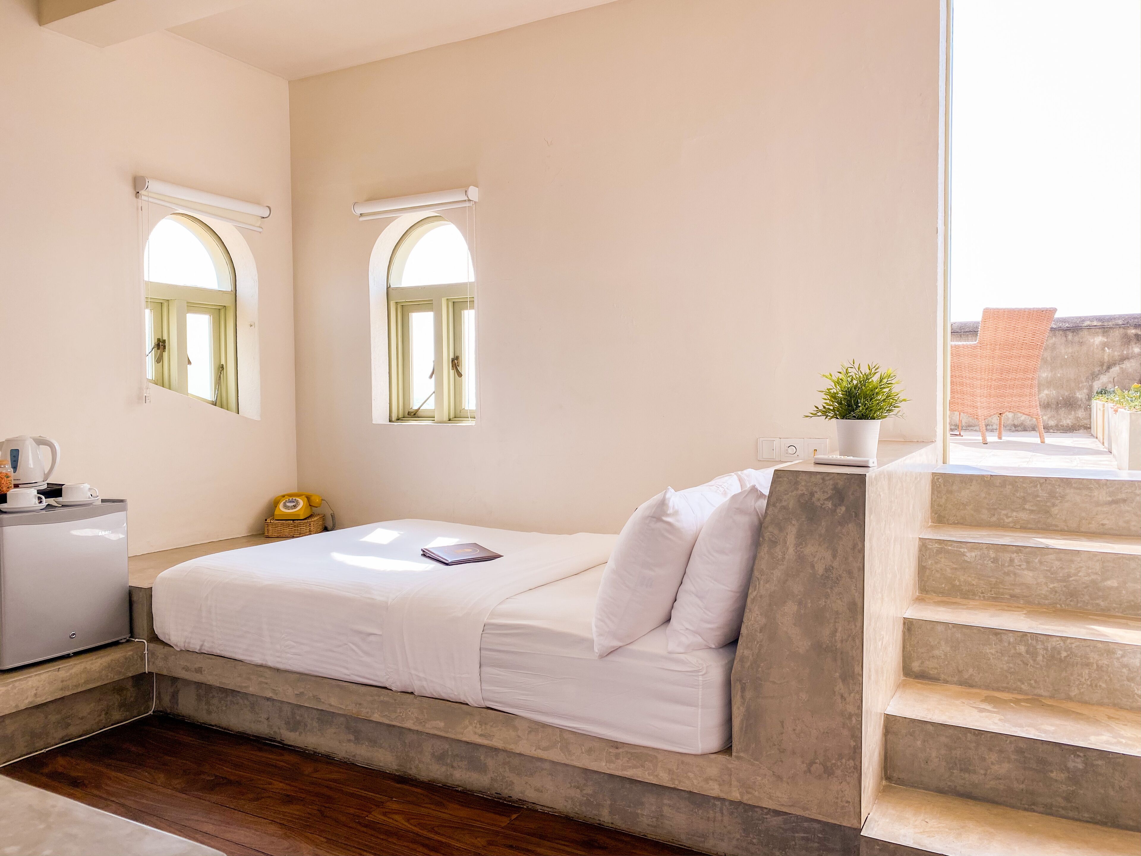 A bedroom with a bed, a desk, a potted plant, and a view of the outside.
