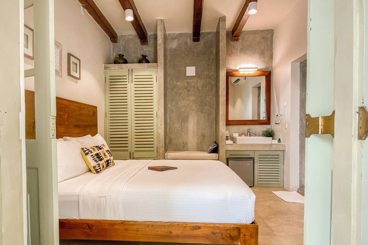 Una camera da letto con un letto in legno, un comodino, un armadio a muro, un bagno con lavandino e specchio, e una luce a soffitto.