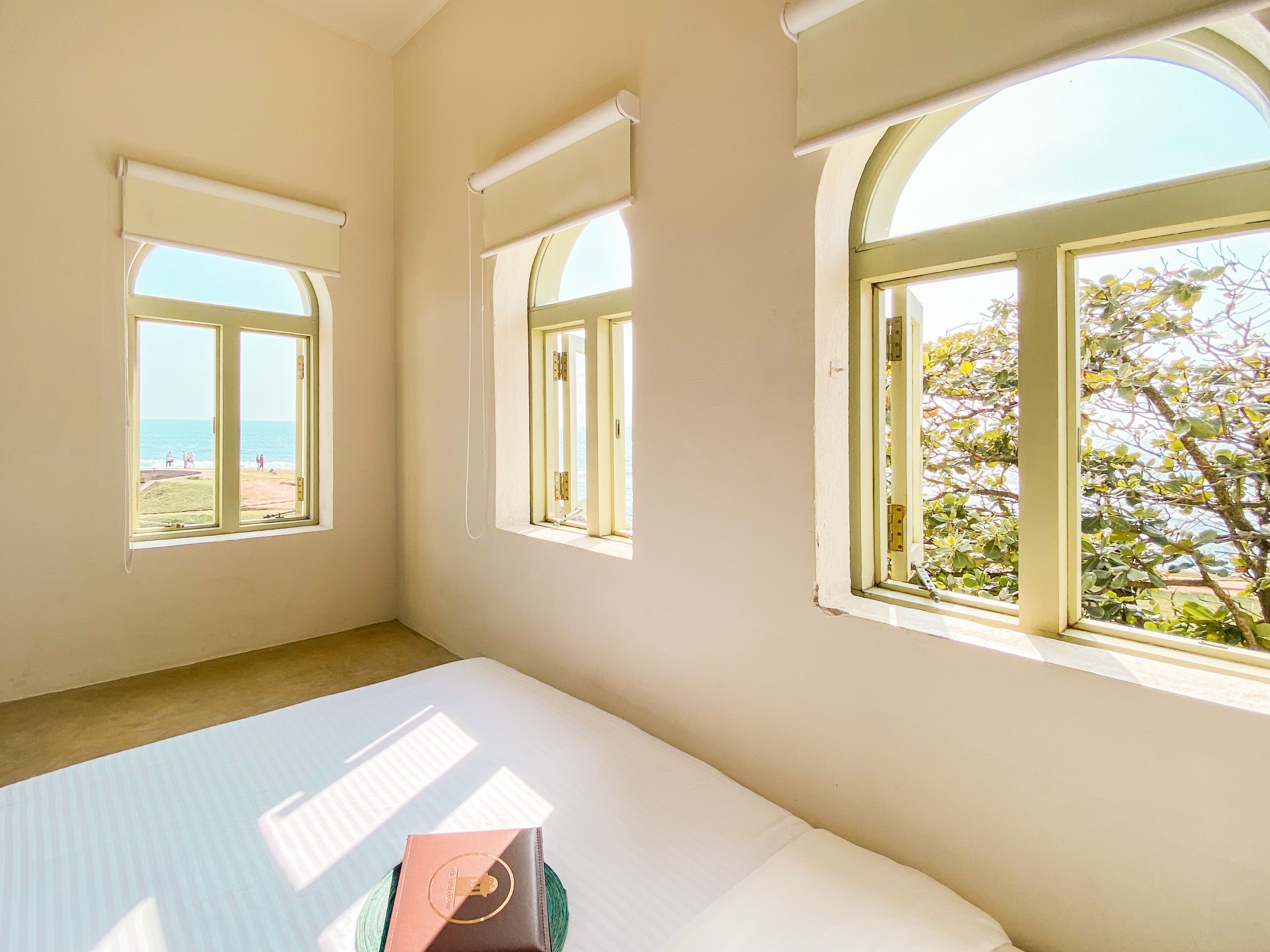 A room with three windows, each with a view of the beach and a tree outside.