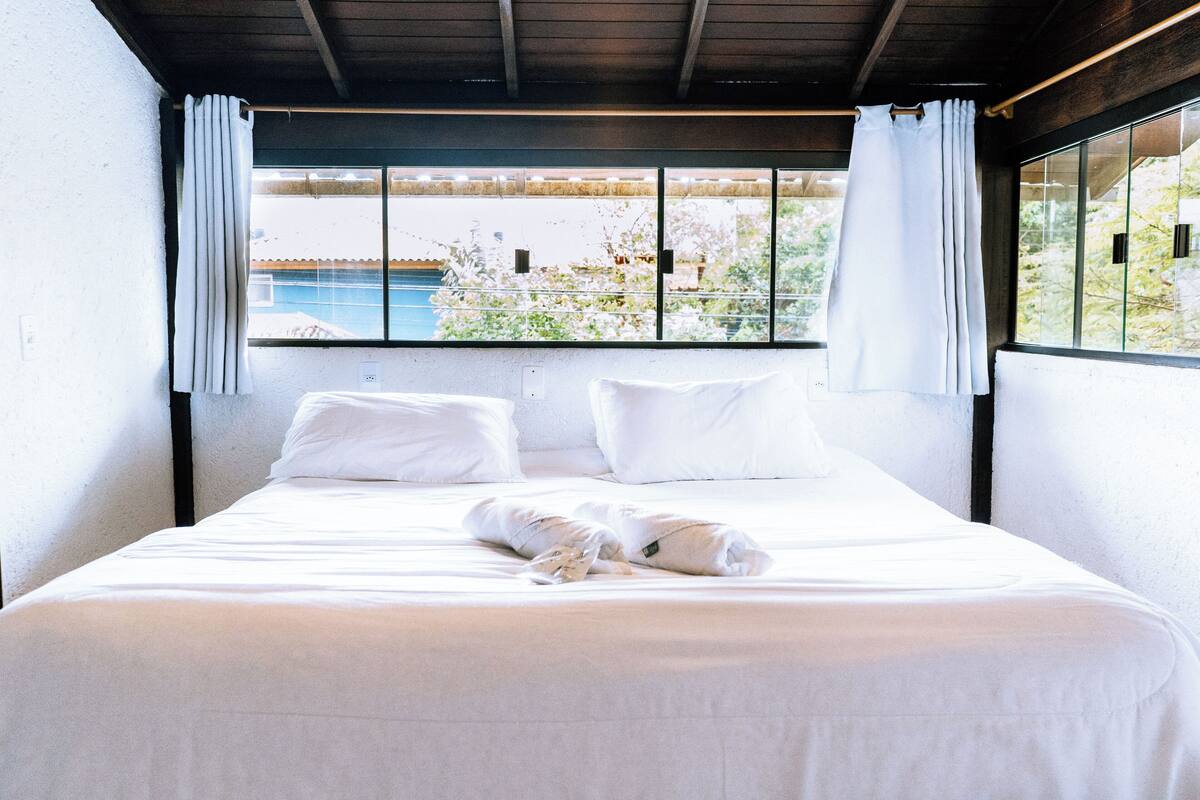 Un lit bien fait, avec du linge de lit blanc et des oreillers, une fenêtre donnant sur une piscine et sur la verdure, et un plafond en bois.