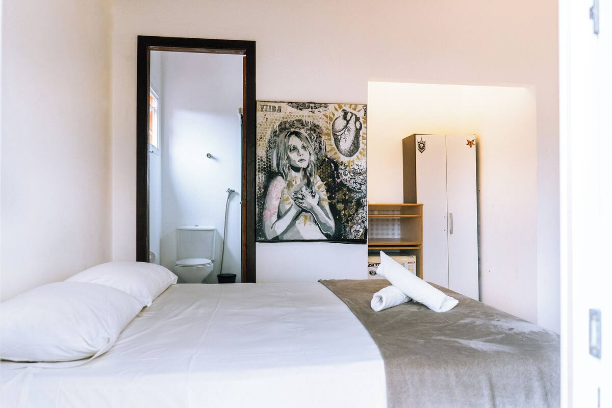 Une chambre à coucher avec un lit, un tableau, une armoire et une salle de bain visible à travers une porte ouverte.