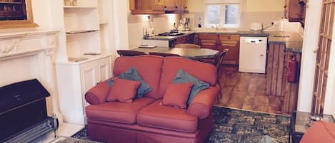 A cozy living room with a red sofa, a glass coffee table, and a fireplace. The kitchen is visible in the background with wooden cabinets and a window.