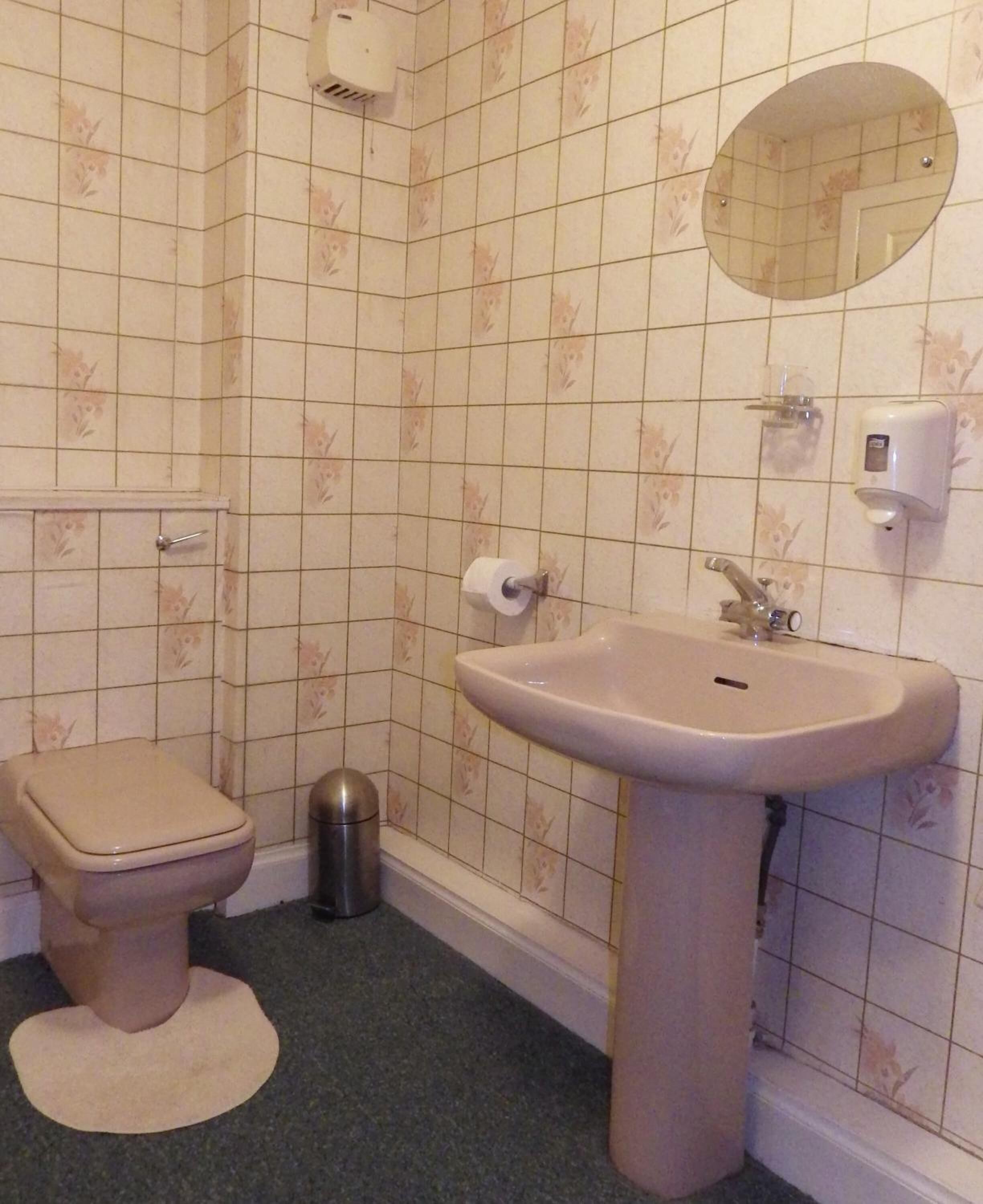 Baño con inodoro, lavamanos, espejo y dispensador de papel higiénico.