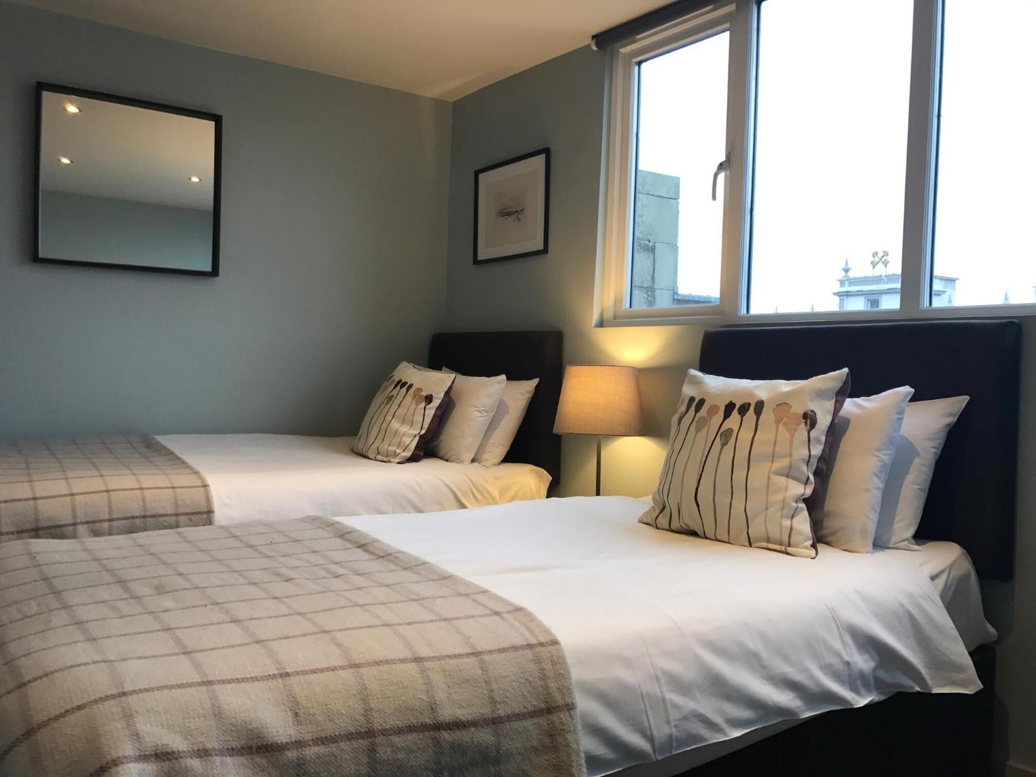 Een hotelkamer met twee bedden, een groot raam, een spiegel en een lijst met een foto aan de muur.