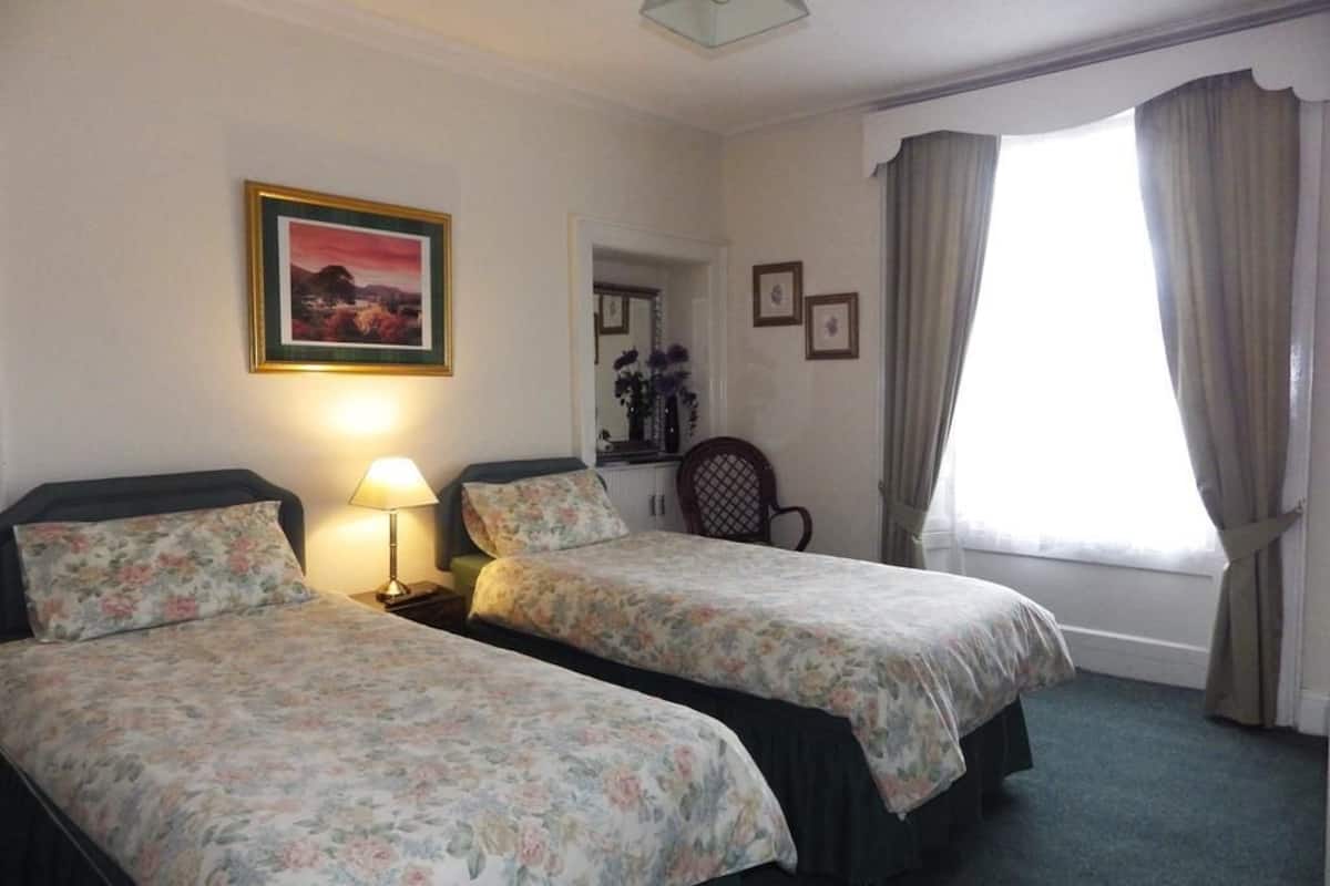 A hotel room with two single beds, a nightstand with a lamp, a framed picture on the wall, a chair, and a window with curtains.
