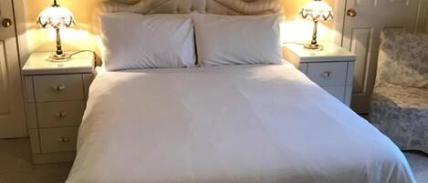 Una cama bien hecha con sábanas blancas, dos mesitas de noche con lámparas, un cuadro enmarcado sobre la cama y una puerta a la derecha.