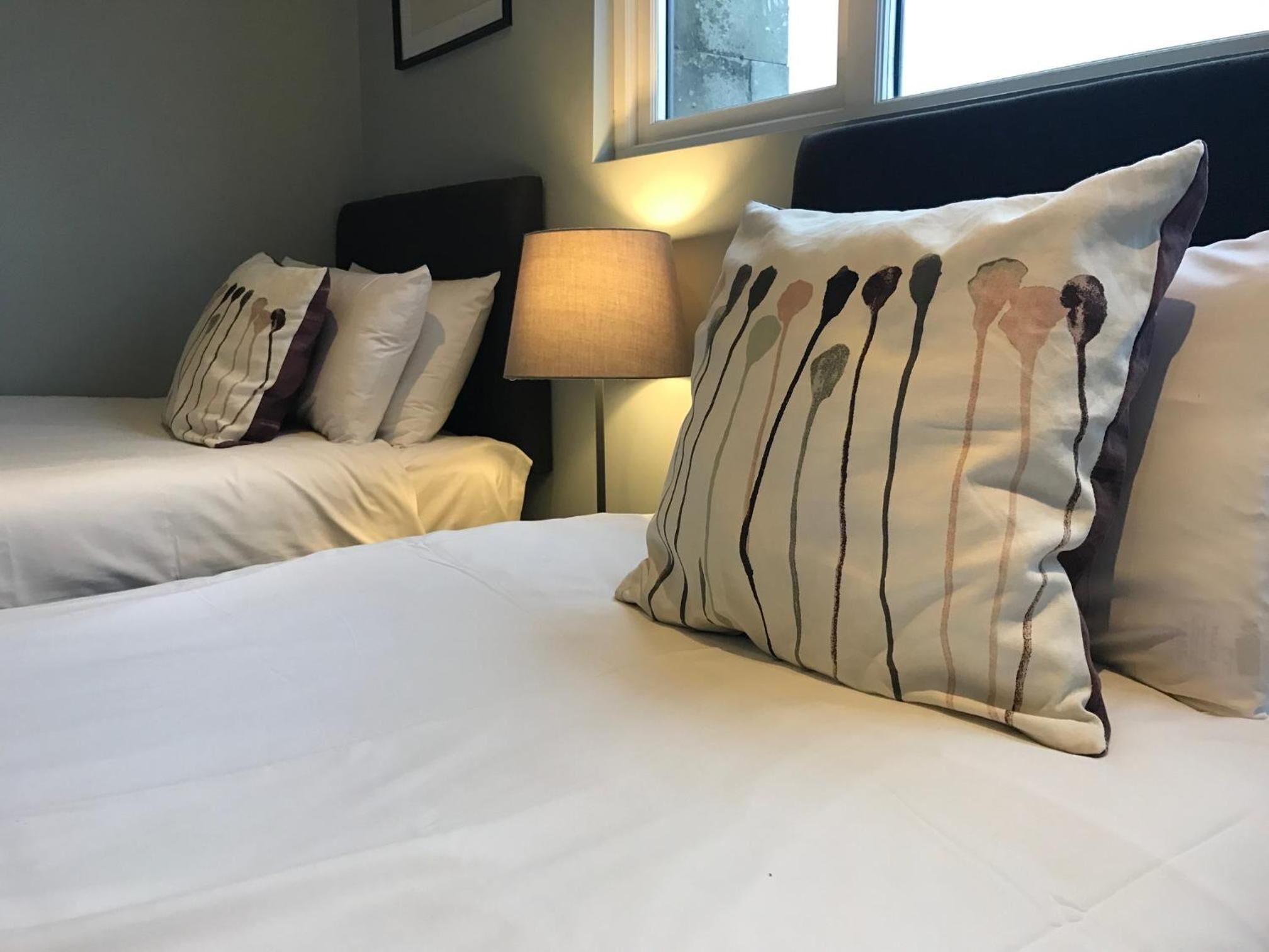 A hotel room with two beds, each with white bedding and striped pillows. There is a lamp on the nightstand between the beds.
