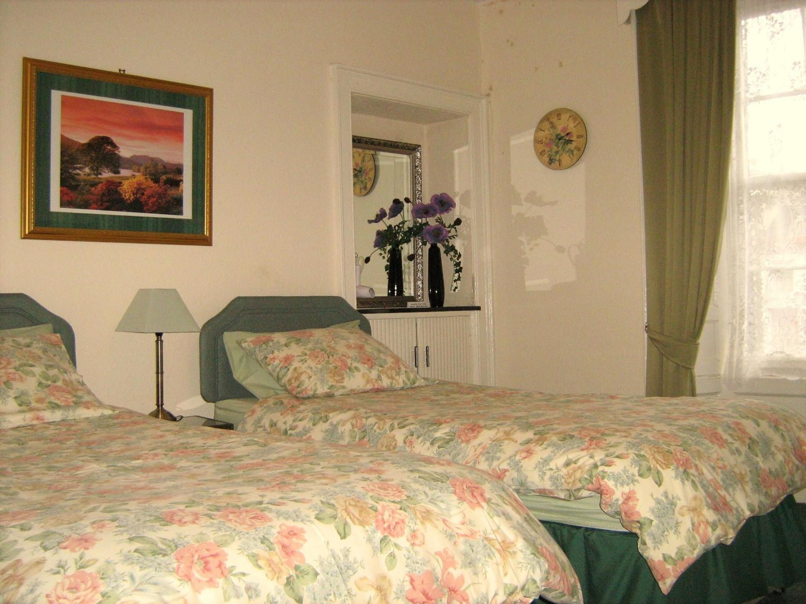 A bedroom with two beds, a nightstand, a lamp, a mirror, a clock, and a window with curtains.