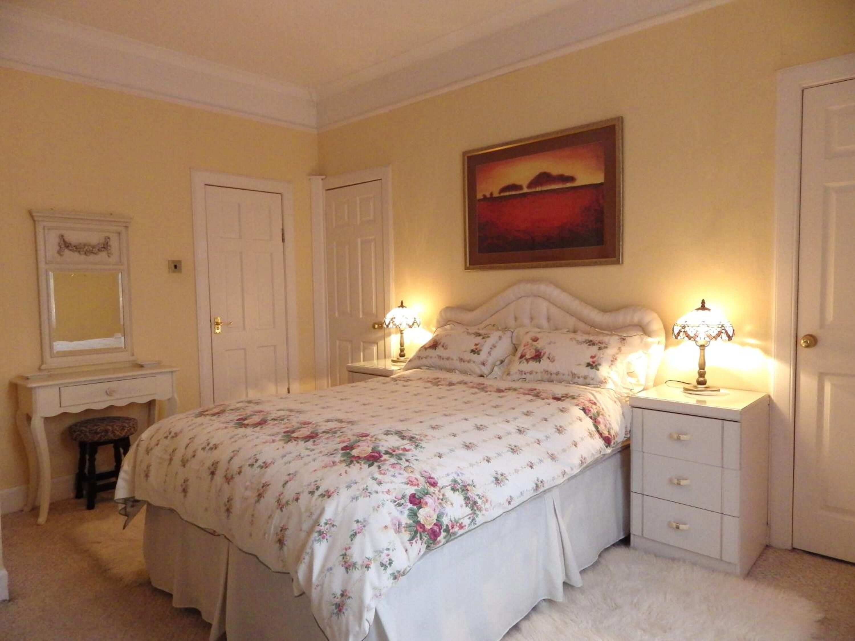 A bedroom with a floral bedspread, two bedside lamps, a nightstand, a dresser, a mirror, and a painting on the wall.