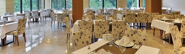 Un comedor amplio con varias mesas y sillas, ventanas grandes que ofrecen vistas a la vegetación exterior.