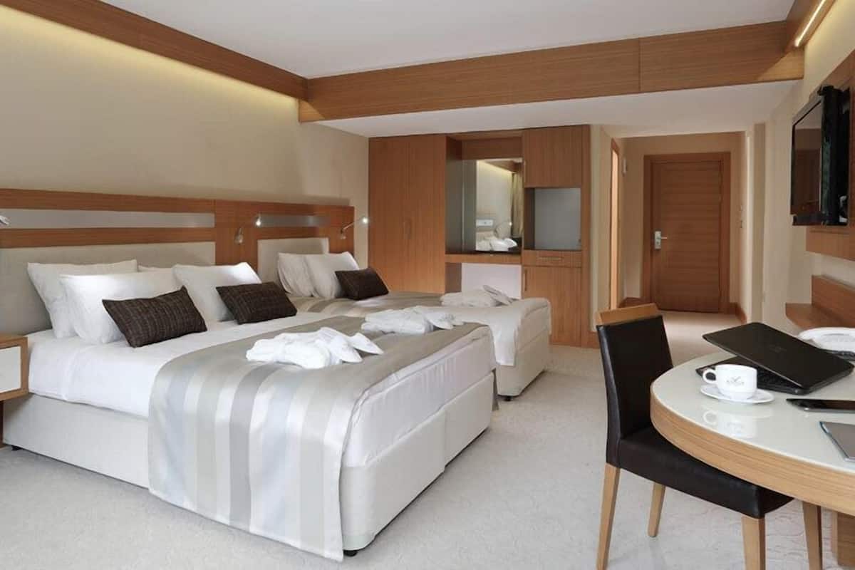 A modern hotel room with a large bed, a bedside table, a chair, and a desk with a laptop and a cup.
