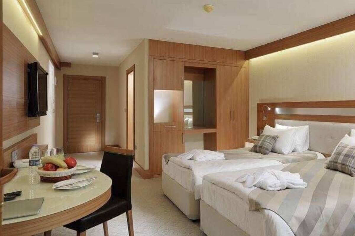 A hotel room with a bed, a desk, and a chair. There is a small table with a fruit bowl and a bottle. The room has wooden walls and a ceiling light.