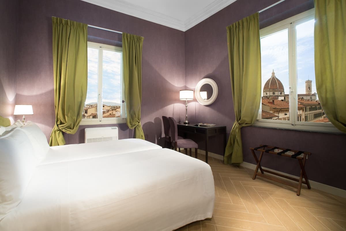 A hotel room with a large bed, two windows with green curtains, a desk with a lamp, and a view of a dome and tower.